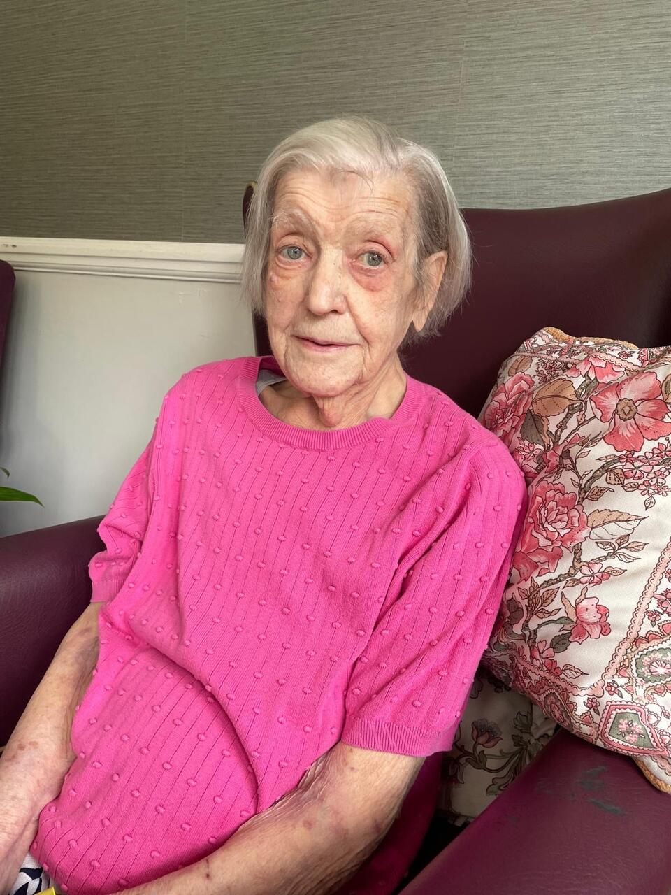 A woman with short grey hair wearing a pink top sat on a burgundy armchair