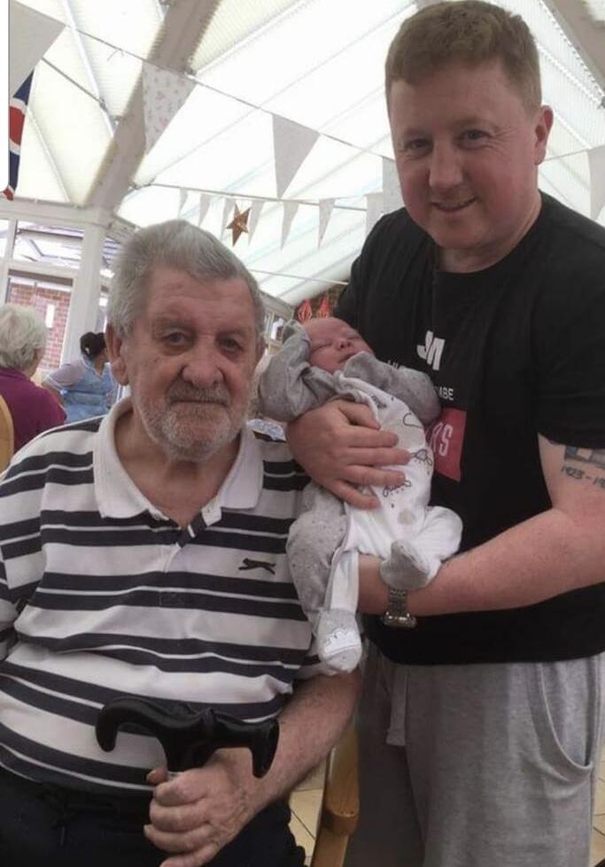 An older man with grey hair and grey beard, stripy t shirt and holding a walking stick and a younger man with light brown hair, wearing a black shirt and holding a baby in a white babygrow