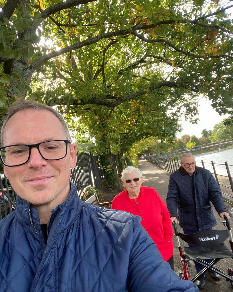 A man wearing glasses and a blue jacket taking a selfie with a woman with short white hair, sunglasses and red jacket and a man wearing a dark jacket using a walker