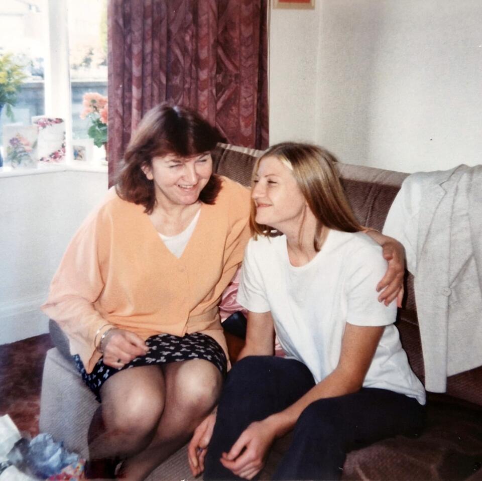 A woman with brown hair wearing an orange jumper and patterned skirt hugging a woman with long blonde hair, white t-shirt and dark trousers while sat on a brown sofa