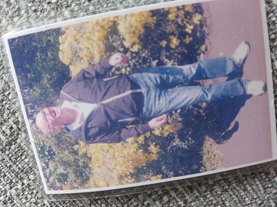A printed photo of a man stood in front of some flowery bushes