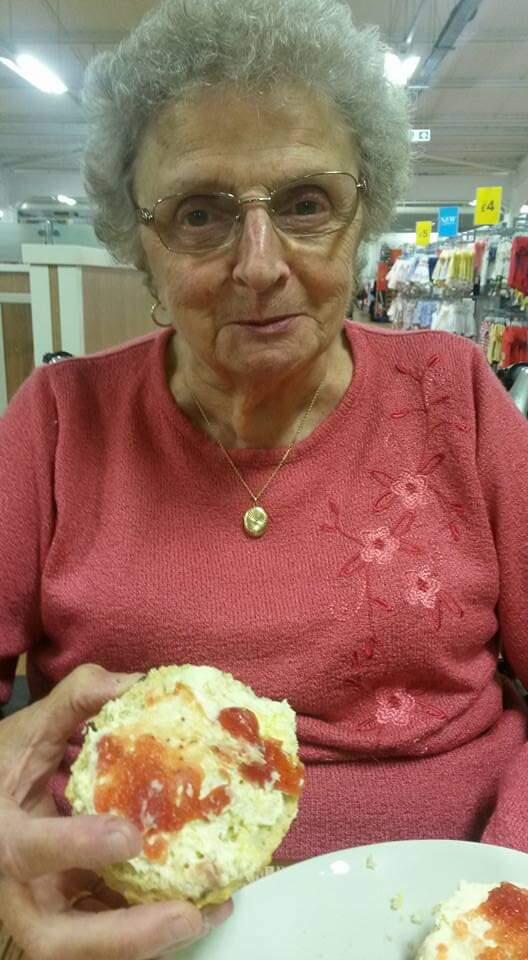 A woman with short, grey curly hair wearing glasses and a pink jumper eating a scone