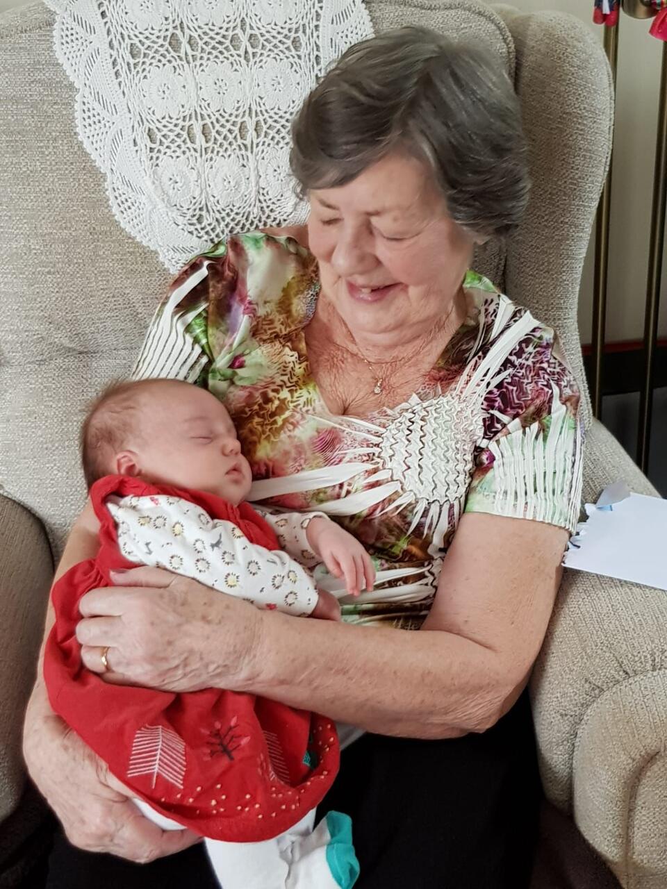 A older woman with short grey hair, wearing a flowery top. She's sat in an armchair and holding a baby