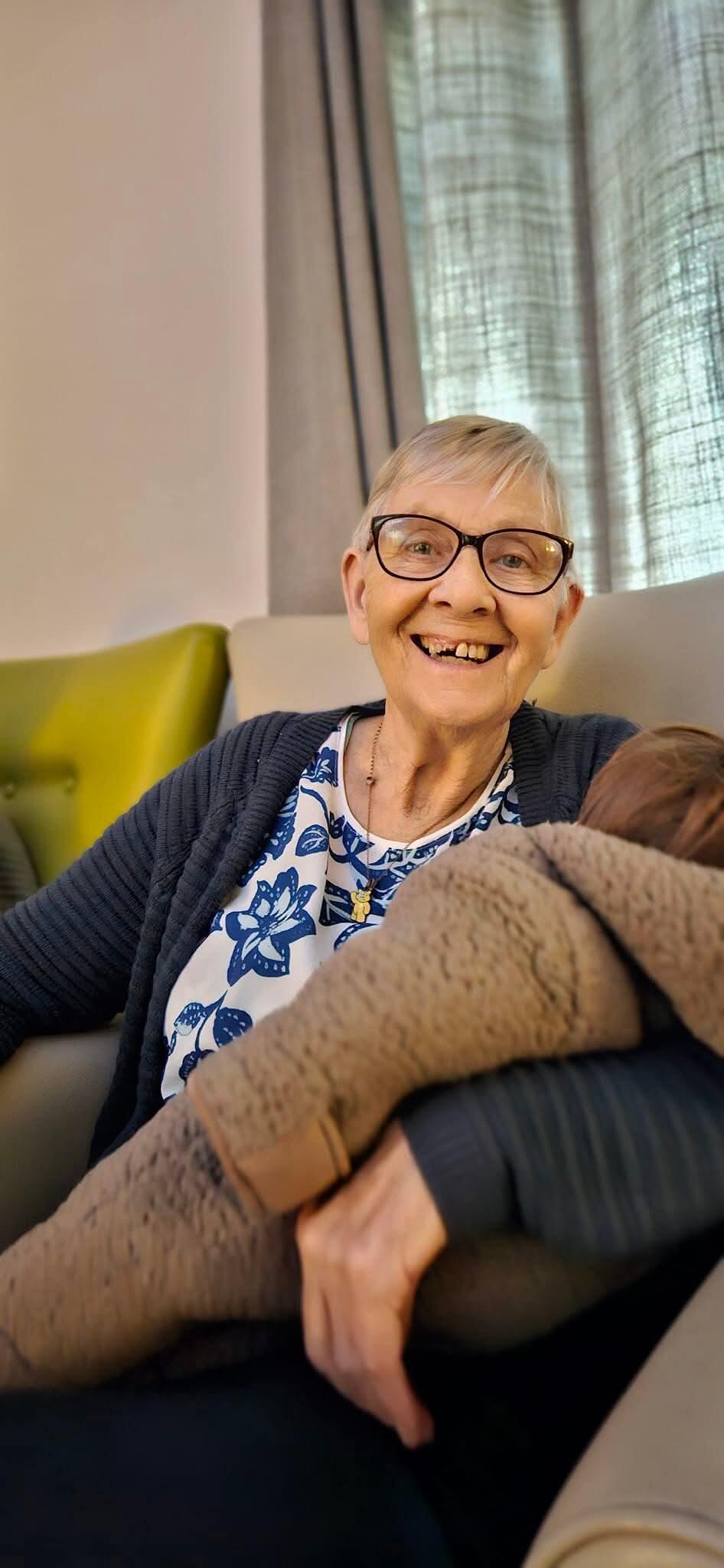 A woman with short grey hair, glasses, patterned top and navy cardigan smiling and sat on a sofa