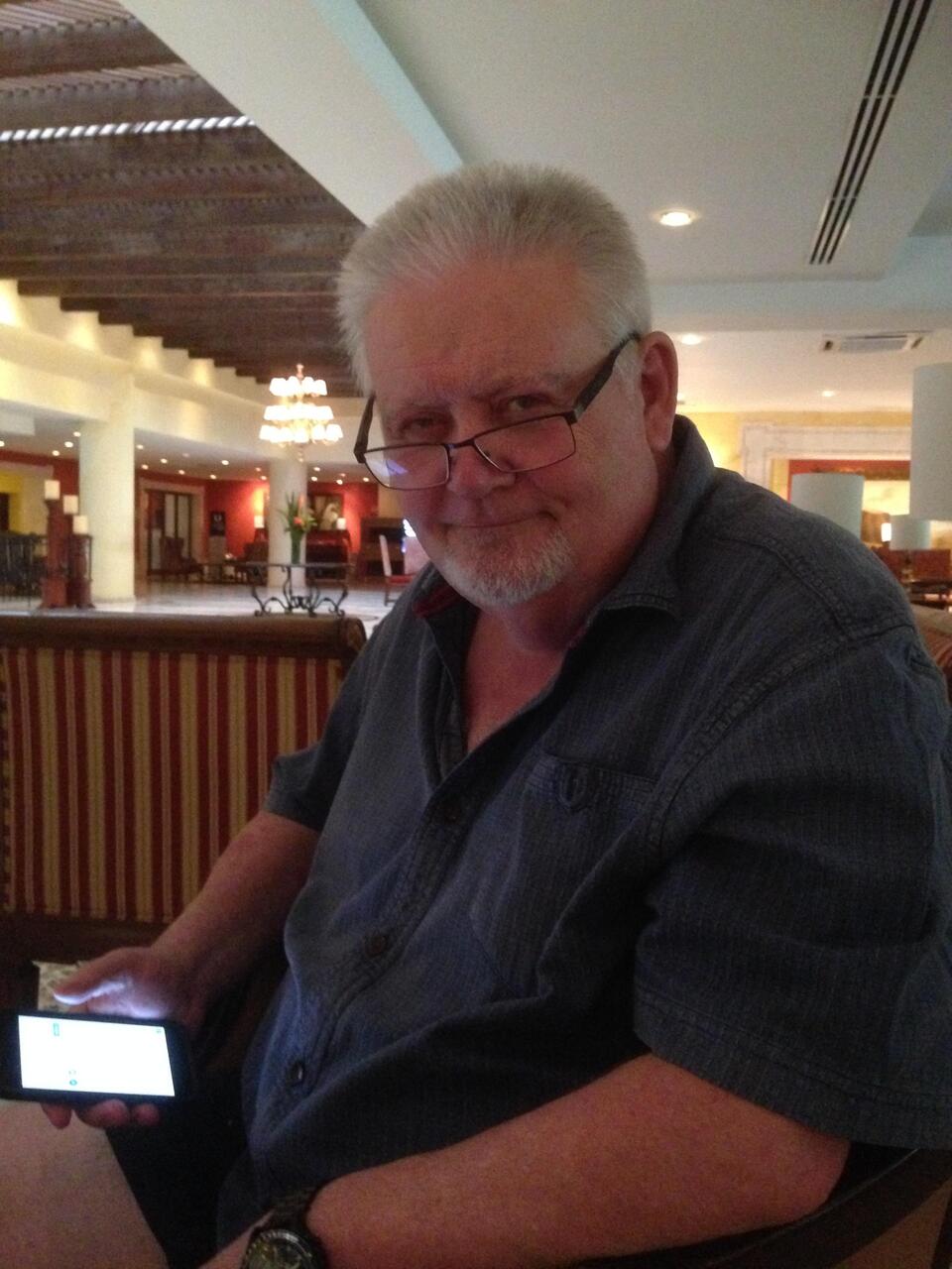 A man with white hair, glasses and a blue shirt holding a phone