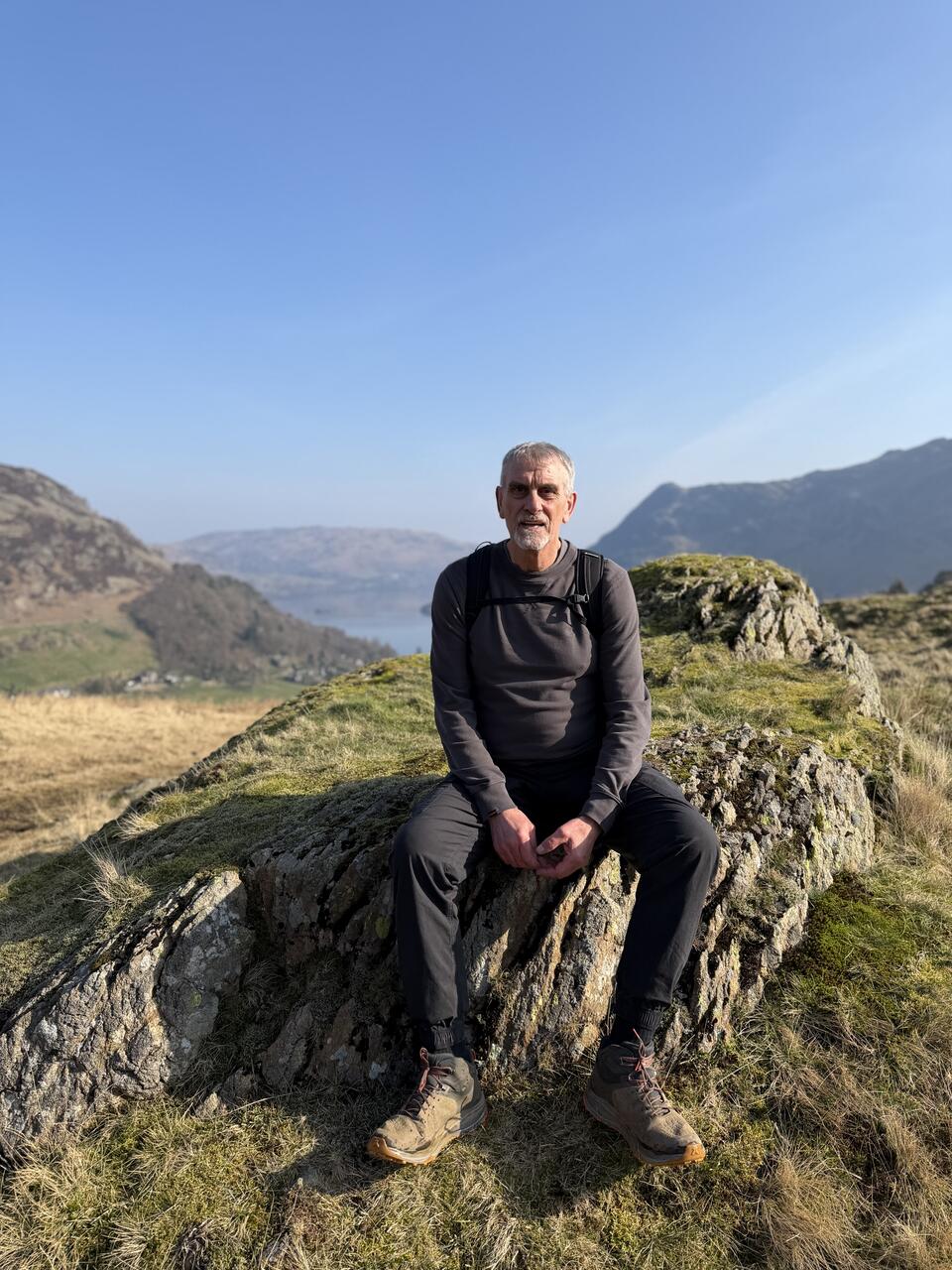 A man wearing a dark top and trousers and glasses with light hair sat on a grassy rock surrounded by hills