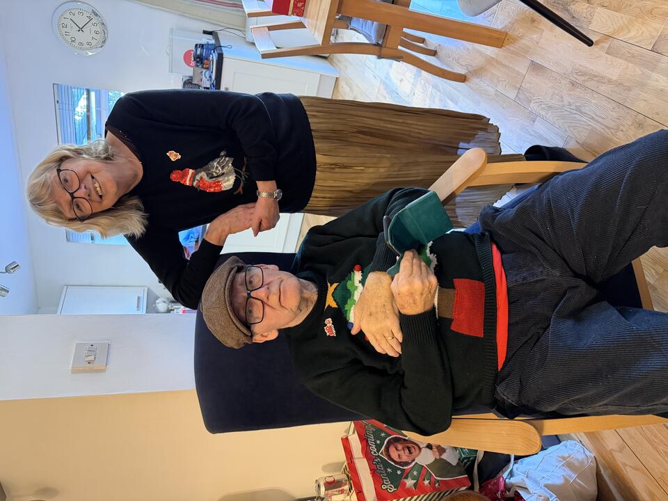 A man sat in a chair wearing a brown hat, glasses, dark jumper and trousers and a woman with a light bob, glasses, dark jumper and gold skirt leaning on the chair