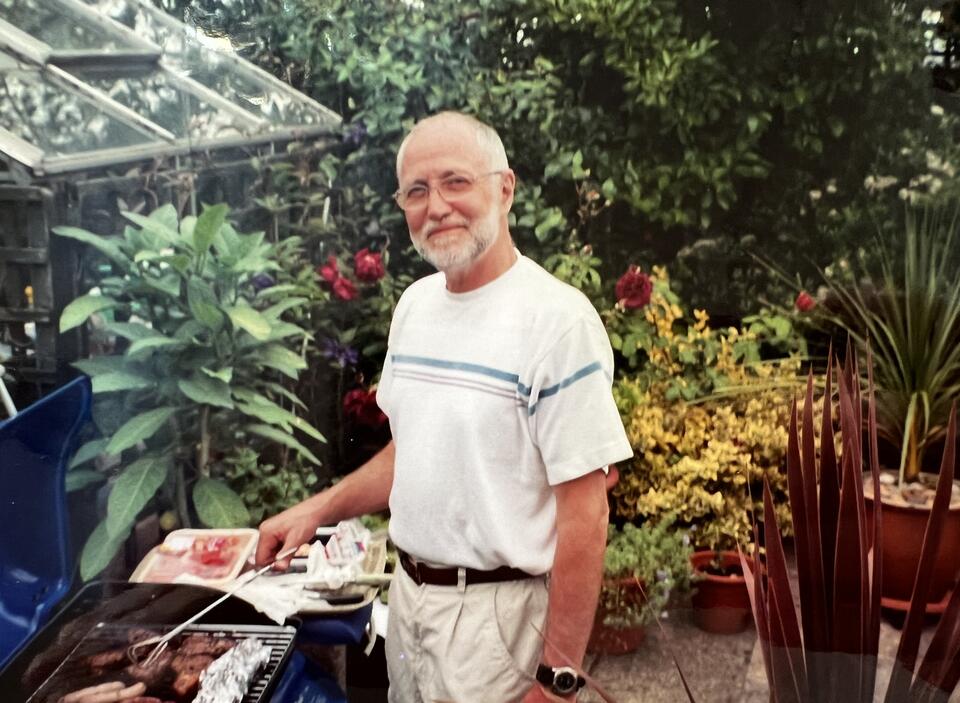 A man with a white beard, white t shirt and beige trousers in a garden