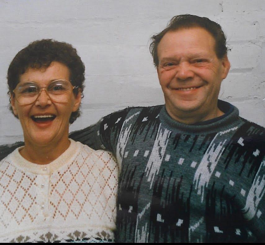 A smiling couple, a lady on the left has short brown hair, glasses and a white cardigan, the man on the right has short brown hair and a green patterned jumper