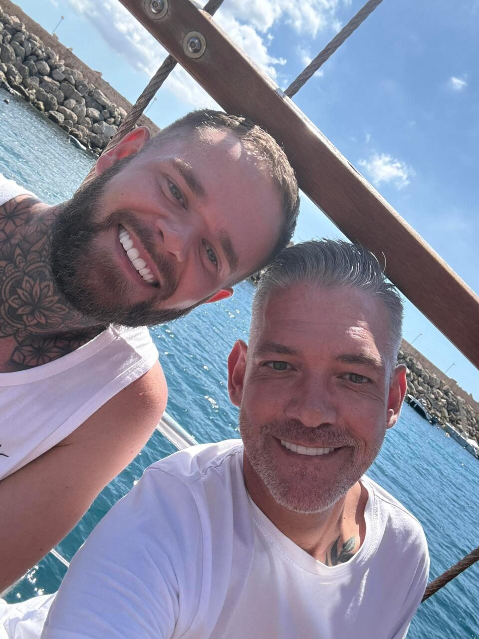 A man with brown hair, beard, a neck tattoo and white vest with another man with grey hair and stubble wearing a white t-shirt