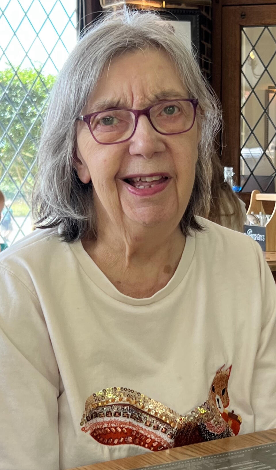 A woman with grey hair, glasses and white top with a glittery motif