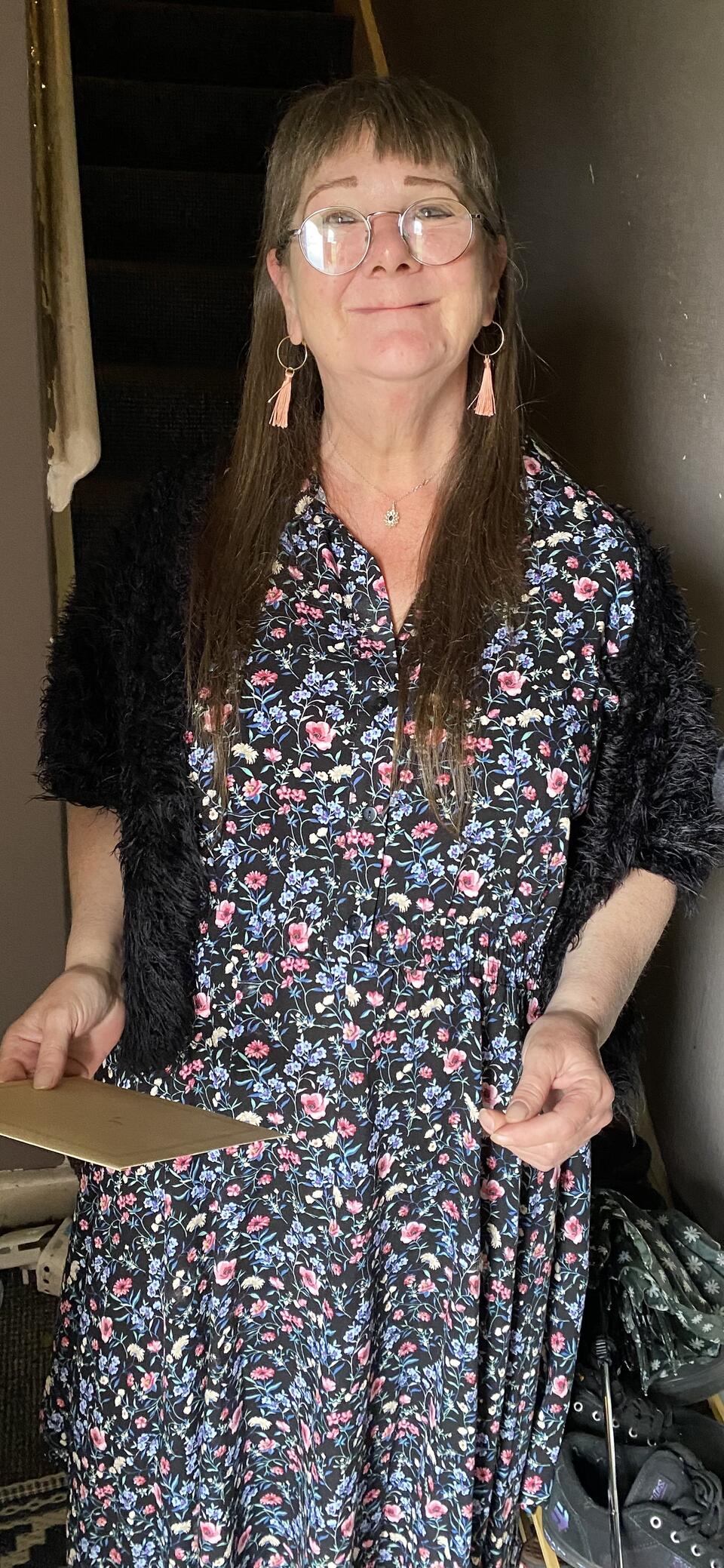 A woman with long brown hair and glasses, long blue patterned dress and dark cardigan