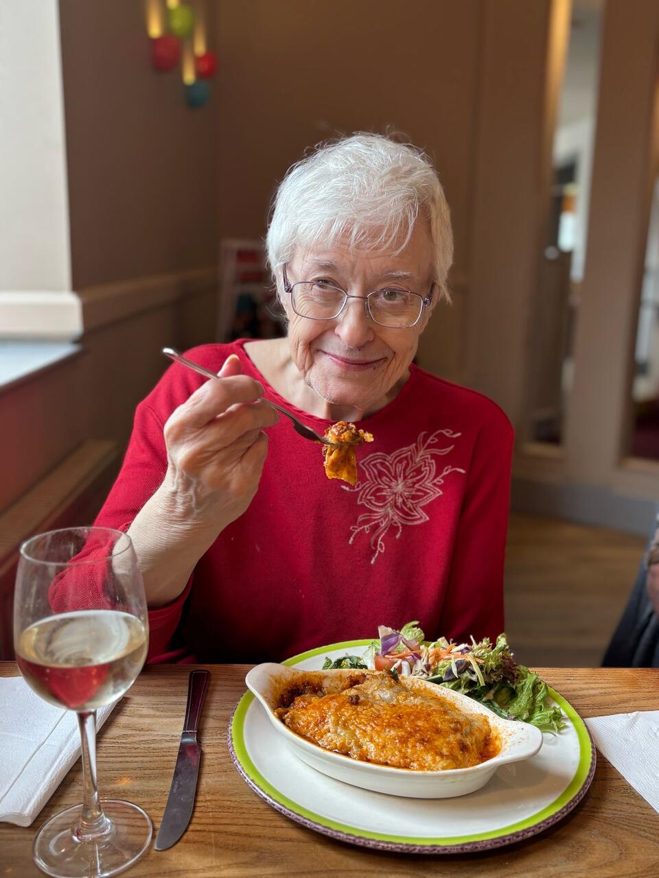 An older woman with short white hair, glasses and wearing a red top eating a lasagna