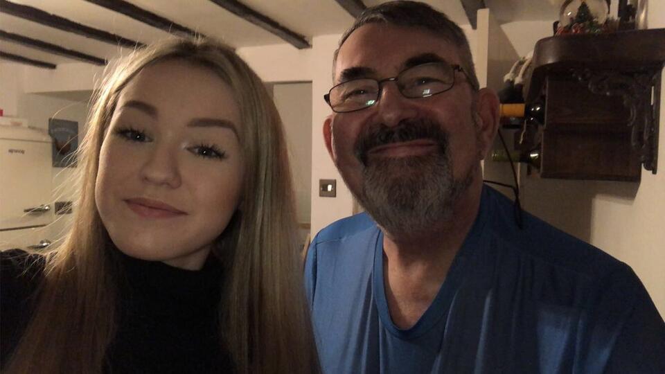 A woman witth long blonde hair taking a selfie with a man with dark hair and a beard wearing glasses and a blue top