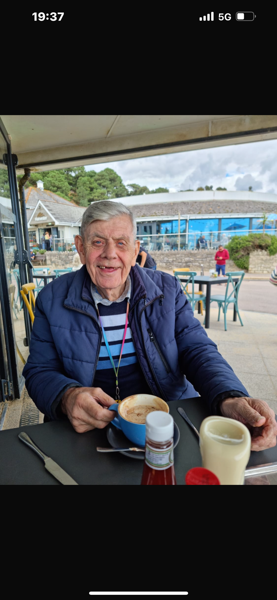An older man with grey hair, wearing a blue jumper and gilet, drinking a milky coffee