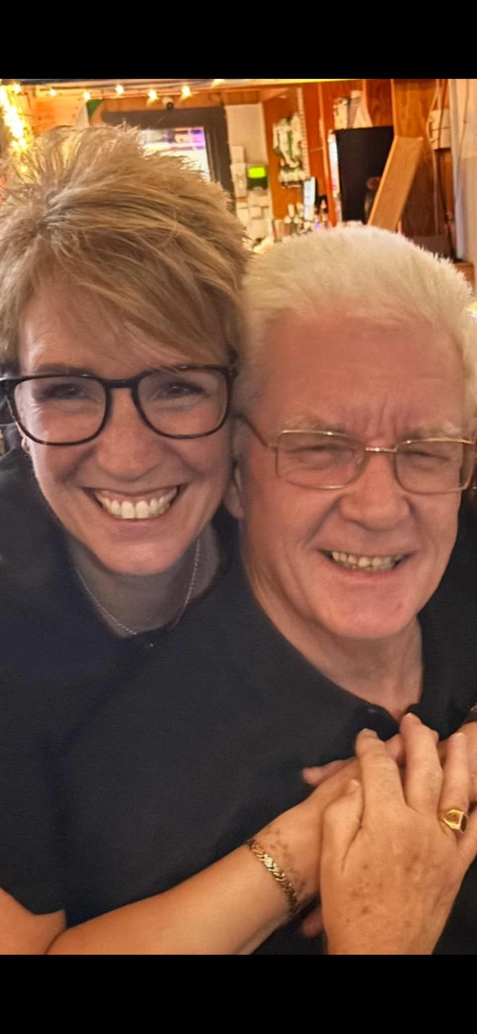 A woman with short blonde hair and glasses hugging an older man with white hair and glasses