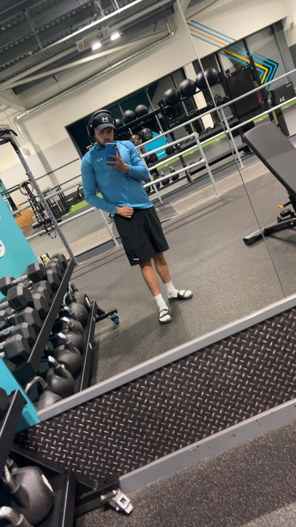 A man wearing a cap, a blue top and shorts taking a selfie in a gym mirror