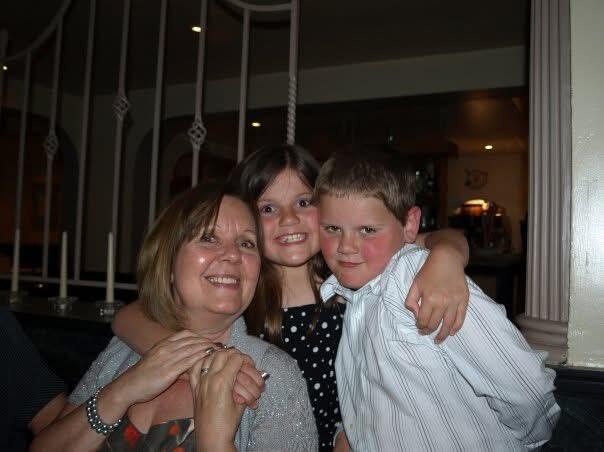A girl with brown hair and a dark dress hugging a woman with brown hair and grey cardigan and a boy with brown hair and white shirt