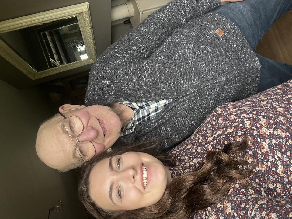 A bald man with glasses wearing a grey cardigan and jeans take a selfie with a woman with long brown hair and a patterned shirt
