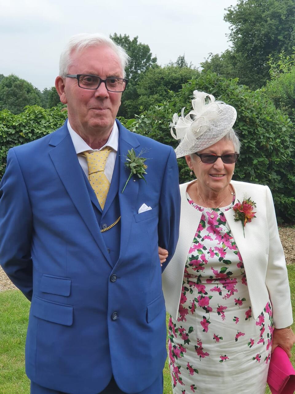 A man with white hair and glasses, wearing a blue suit with a white tie and a woman with a white hat and blazer and white and pink floral dress