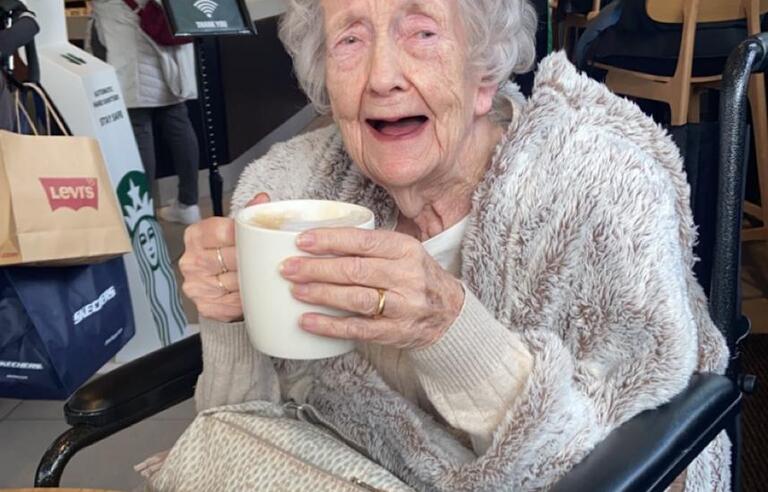 A woman with short grey hair wearing a grey top drinking a warm drink in a cafe