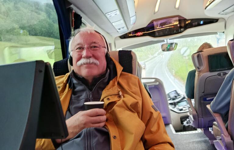 A man with white hair and moustache wearing a yellow coat and drinking a coffee on a coach