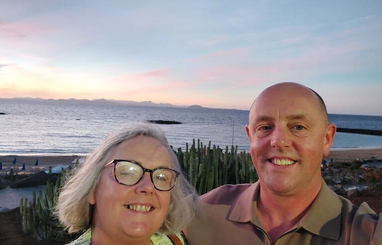 A woman with short grey hair and glasses and a bald man with a green top on a beach