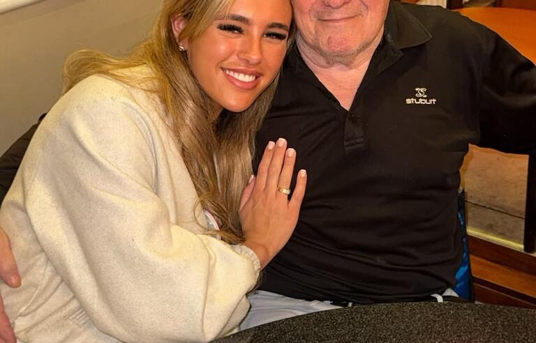 A younger woman with long blonde hair wearing a white jumper hugging an older man with white hair wearing a blue t shirt