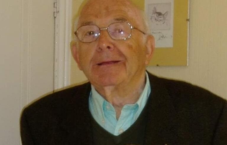 A bald man wearing glasses, light blue top and dark jumper