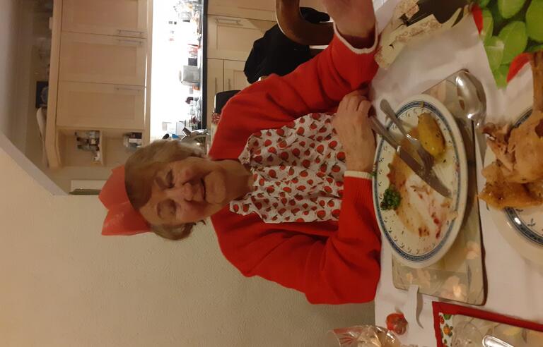 A woman wearing a red patterned top, red cardigan and Christmas cracker hat eating a roast dinner