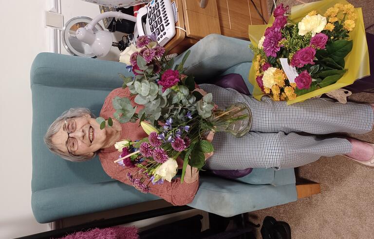 A woman with short grey hair, pink top and grey trousers sat in a green chair holding flowers