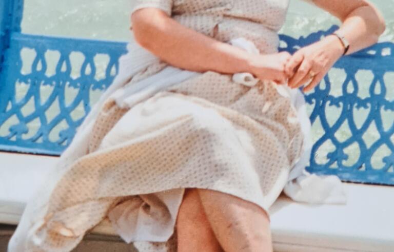 A woman with light hair wearing a cream dress sat on a bench by the sea
