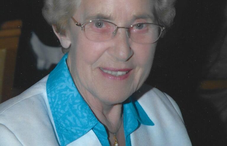 A smiling woman with short, grey hair, wearing glasses and blue shirt and white blazer