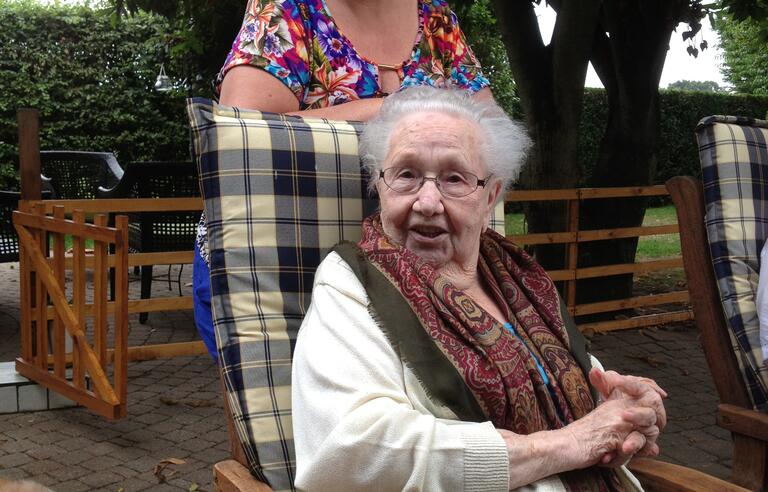 An older woman sat on a chair in a garden with a younger woman stood behind her
