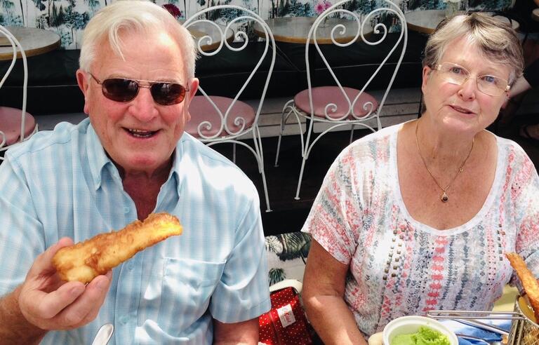 A man with white hair, sunglasses and a light blue shirt holding battered fish and a woman with white and grey hair and patterned top