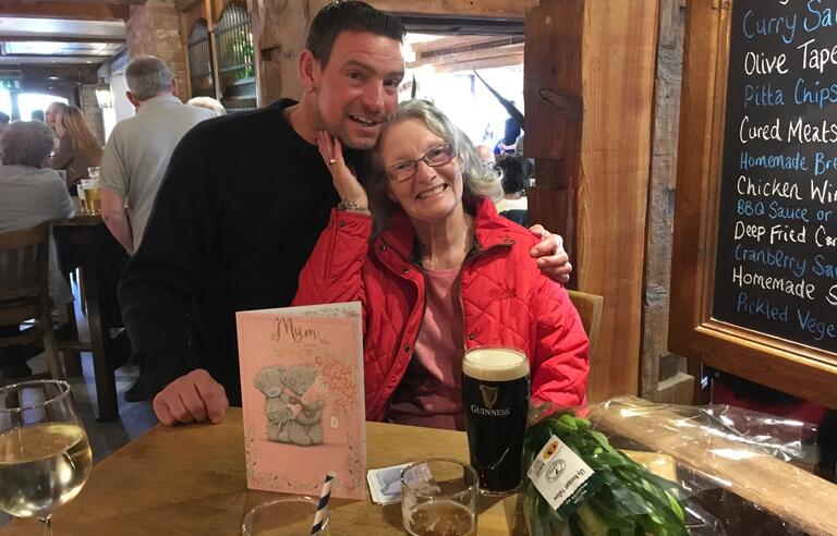 A man with brown hair and dark top and a woman with light wavy hair wearing a red coat in a pub