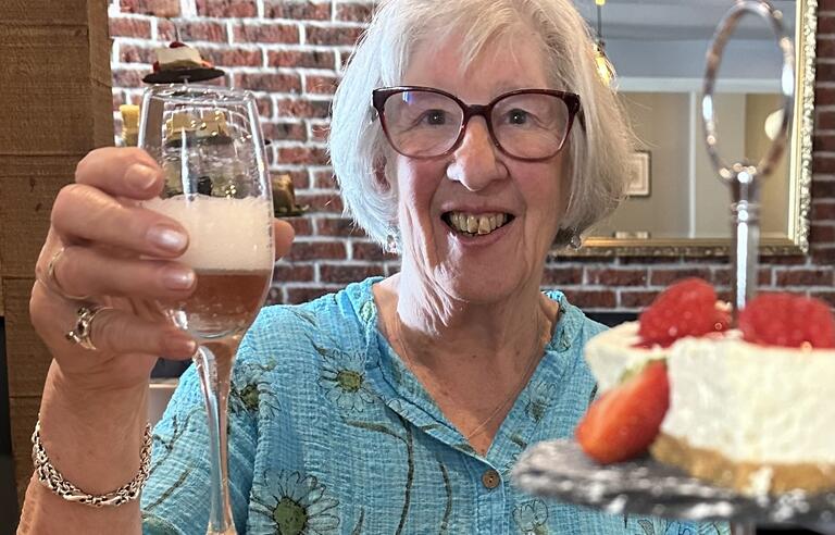 A woman with short white hair, glasses and a blue shirt holding a glass of prosecco