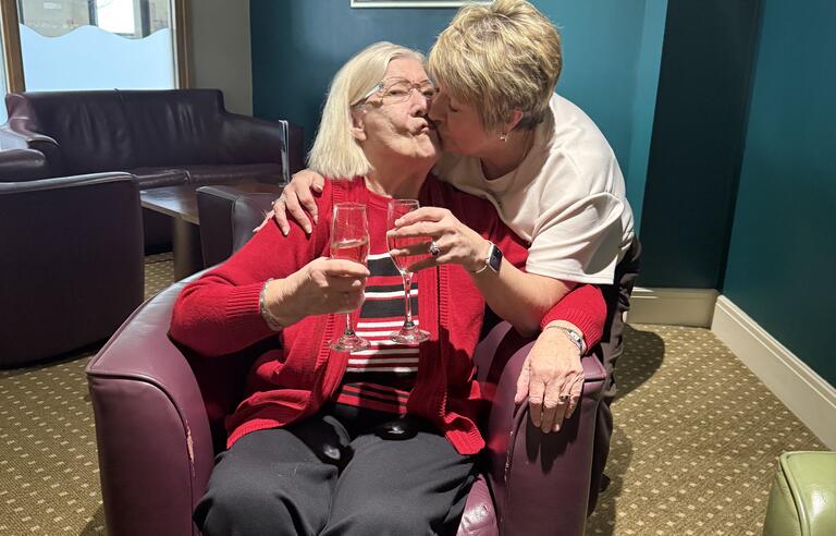 A woman behind a chair kissing a woman sat in the chair