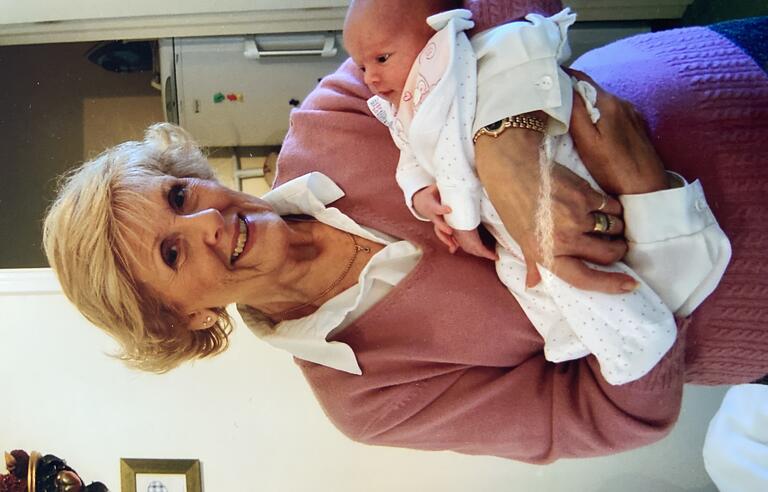 A woman with short blonde hair wearing a white shirt and pink jumper holding a baby dressed in white