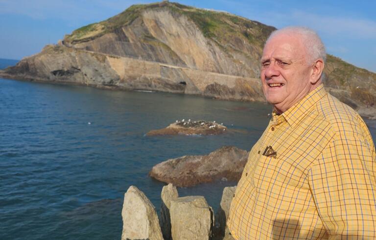 A man wearing a yellow shirt stood in front of sea and a hill