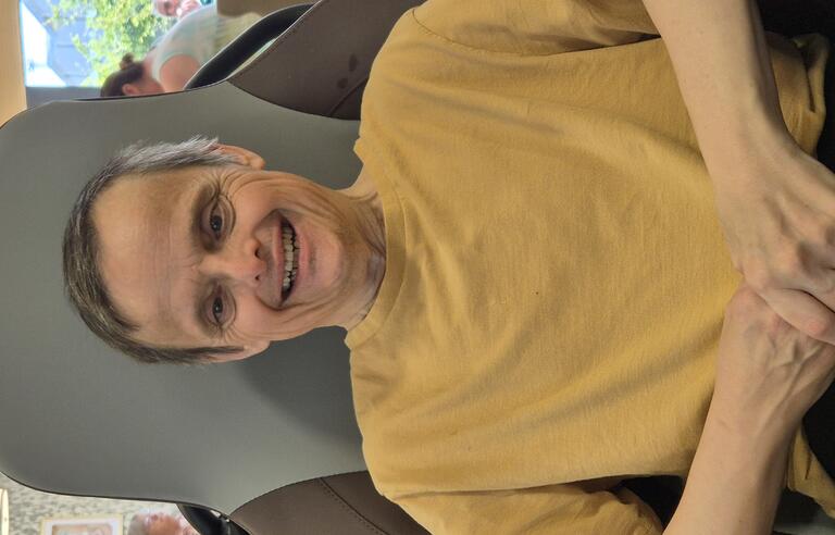 A smiling man wearing a yellow t-shirt sat in a chair