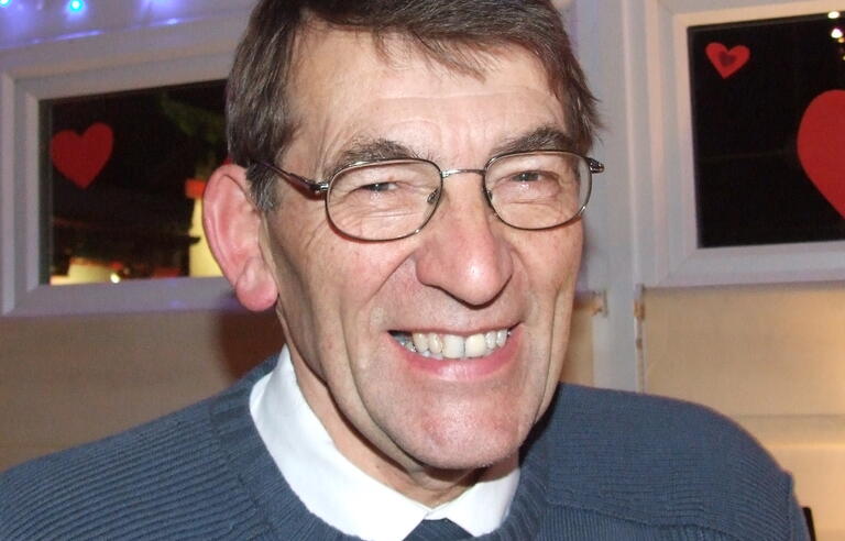 A smiling man with brown hair wearing glasses, a white shirt and blue jumper