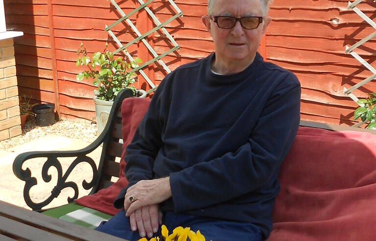 A man with white hair, sunglasses and dark jumper sat on a bench in a garden