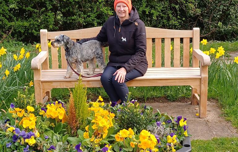 A woman with an orange hat, brown coat and jeans sat on a bench with a small dog