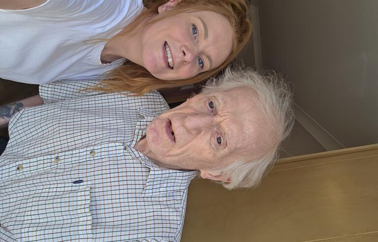 A woman with a ginger ponytail, wearing a white t shirt and a man with white hair wearing a white and green check shirt