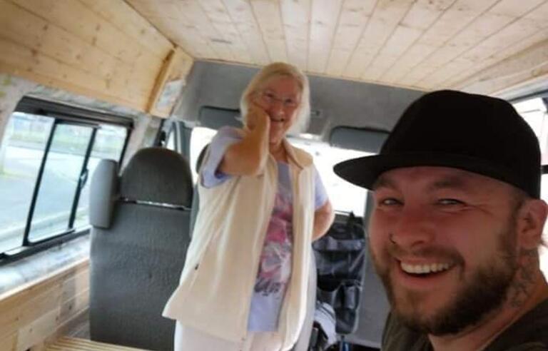 An older woman with short white hair, a white gilet and white trousers and a man with a black cap, beard and black t shirt