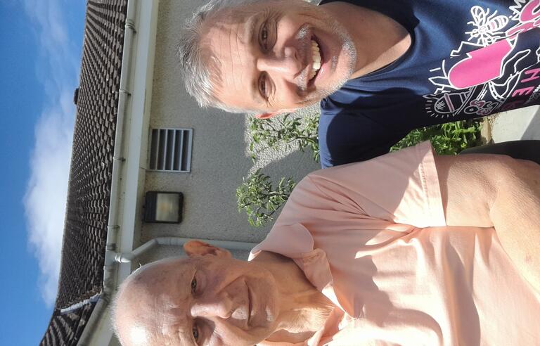 A bald man wearing a peach shirt and a man with grey hair wearing a blue patterned t-shirt in a garden