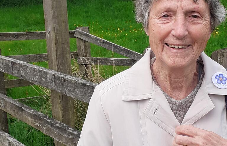 A lady with grey hair smiling. She's wearing a white coat and is stood in front of a sign