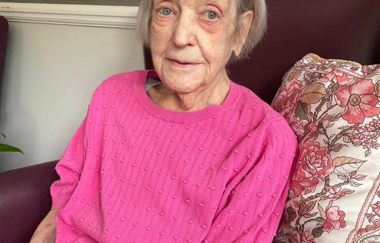 A woman with short grey hair wearing a pink top sat on a burgundy armchair