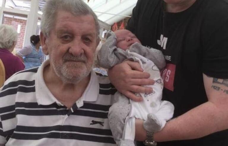 An older man with grey hair and grey beard, stripy t shirt and holding a walking stick and a younger man with light brown hair, wearing a black shirt and holding a baby in a white babygrow